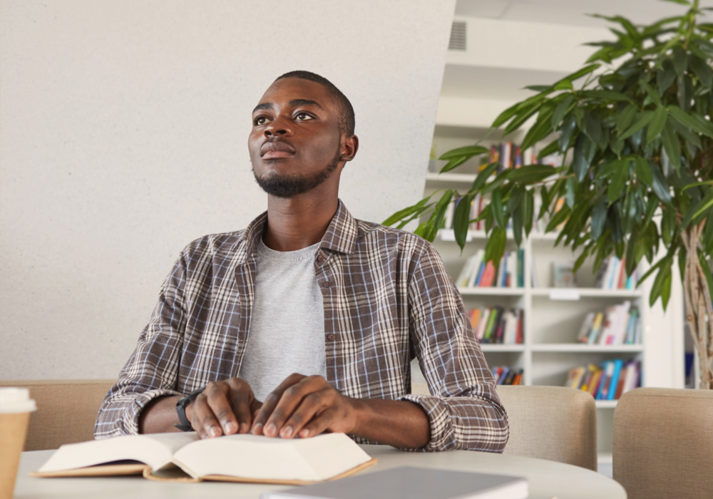 A young man sits at a table reading a heavy book using Braille.