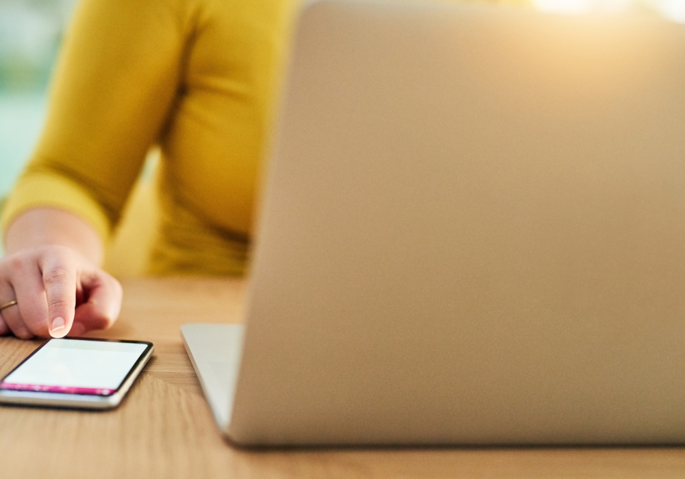 A hand is seen tapping a cell phone screen next to an open laptop.